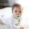 A baby lying on a white rug with a pacifier in its mouth. Produktbild-Vorschau