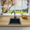 A black kitchen sink and faucet are shown on a wooden countertop, with a window view of a scenic landscape in the background. A bowl of oranges is placed to the right of the sink. Produktbild-Vorschau