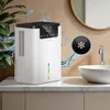 A white dehumidifier is shown on a countertop in a bathroom next to a sink and a faucet, with water splashes around it. Náhled obrázku produktu