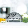 A stainless steel steamer basket is placed on a table with cut vegetables and cauliflower in the background. Produktbild-Vorschau