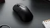 A black wireless mouse is placed on a dark gray surface, with a small calendar visible on the right. Voir les images du produit