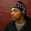 A man wearing headphones and a beanie stands against a red wall. Produktbild-Vorschau