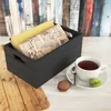 A dark wooden crate filled with books and newspapers, placed on a wooden surface next to a cup of tea and macarons. Anteprima dell’immagine prodotto