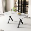 A white marble-patterned dining table with black X-shaped legs is shown in a well-lit room with a bookshelf in the background. Náhľad obrázku produktu