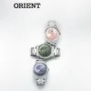 Three Orient watches are stacked on a white background. The top watch has a pink face, the middle watch has a green face, and the bottom watch has a blue face. The Orient logo is visible on each watch face. The brand name "ORIENT" is also displayed in larg Produktbild-Vorschau