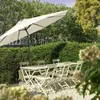 An outdoor patio set with a large umbrella, table, and chairs, surrounded by lush green hedges and trees. Produktbild-Vorschau
