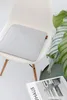 A light gray seat cushion with a wooden tag is placed on a white chair. A plant and a magazine are on the floor next to the chair. Produktbild-Vorschau