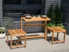 A wooden table and two stools are set up on a patio next to a brick wall and some greenery. Produktbild-Vorschau