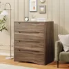 A dresser with four drawers is shown in a room with a floor lamp and framed pictures on the wall. Náhled obrázku produktu