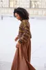 A woman walks outdoors wearing a patterned sweater and a long brown skirt. Produktbild-Vorschau