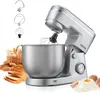 A silver stand mixer with various attachments, a bowl, and baked goods in the background. Produktbild-Vorschau