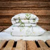 A folded duvet and pillow with a floral pattern are stacked on a white fluffy rug, placed on a wooden pallet, with a rustic wooden wall in the background. Produktbild-Vorschau