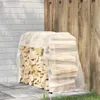 A stack of firewood covered by a transparent tarp, leaning against a wooden fence and a brick wall. Podgląd zdjęcia produktu