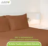 A close-up view of a bed with brown sheets and pillows, with a bedside lamp and a wall lamp in the background. Produktbild-Vorschau