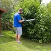 A man is trimming a large bush with a hedge trimmer in a backyard. Produktbild-Vorschau