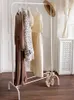 A white metal garment rack with clothes and a hat on it, placed on a wooden floor in front of a white door and a radiator. Náhled obrázku produktu