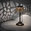 A solar-powered table lamp with a decorative metal shade casts intricate shadows on a wall and surface. Produktbild-Vorschau