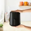 Hands holding a black air fryer on a wooden countertop with books and mugs in the background. Produktbild-Vorschau