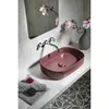 A maroon red oval vessel sink is shown on a light gray countertop in a modern bathroom setting. The sink is accented by a chrome faucet and a small decorative item. The background features a concrete-like wall and a mirror reflecting pink flowers in a vase Produktbild-Vorschau