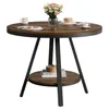 A round dining table with a wooden top and black metal legs, set against a white background. It has a lower shelf with books and a decorative item. A vase of flowers, a mug, and a plate with pancakes are on the tabletop. Produktbild-Vorschau