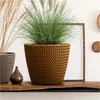 A brown knitted planter with a green grass plant inside, placed on a wooden shelf next to a smaller grey planter and a brown vase. A wooden frame is in the background. Podgląd zdjęcia produktu