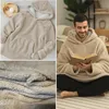 A collage of images showing a beige sherpa hoodie blanket, a close-up of its texture, and a man wearing it while reading a book. Produktbild-Vorschau