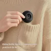 A hand holding a small, black, round electronic device with a digital display, placing it into the pocket of a beige knitted sweater. Produktbild-Vorschau