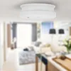 A white smoke detector is mounted on a white ceiling, with a blurred living room visible in the background. Produktbild-Vorschau