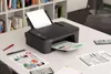 A Canon printer is shown on a desk with papers and pens, with a bookshelf in the background. Produktbild-Vorschau