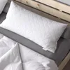 A white quilted pillow on a bed with white bedding and a gray bolster pillow. Náhled obrázku produktu