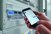 A person is holding a smartphone and interacting with a digital timer device mounted on a control panel. Produktbild-Vorschau