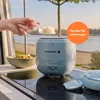 A hand is shown adding rice to a bowl next to a mini rice cooker, with a scenic outdoor background. Produktbild-Vorschau