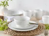 A white porcelain dinnerware set is arranged on a woven placemat, with a vase of greenery in the background. Produktbild-Vorschau