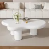 A white, irregularly shaped coffee table with a vase of white flowers and books on it, placed in a modern living room with a white sofa. Produktbild-Vorschau