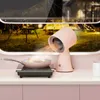 A pink mini range hood is shown on a kitchen counter, directing airflow towards a pan on an induction cooktop. The background shows a campsite scene through a window. Náhľad obrázku produktu