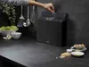 A person's hand is reaching for a set of black cutting boards with steel accents, which are stored in a stand on a dark countertop. Other kitchen utensils and ingredients are visible in the background and foreground. Anteprima dell’immagine prodotto