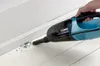 A hand is using a blue and black hand vacuum cleaner to clean up small green beads from a white wooden floor near a white baseboard. Produktbild-Vorschau