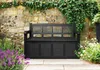 A black storage bench sits in a garden setting against a stone wall covered in ivy, with purple flowers in the foreground and green plants to the right. Produktbild-Vorschau