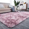 A fluffy pink rug is laid out in a living room setting, in front of a beige sofa. A blanket with pom-poms is draped over the sofa, and a plant is visible in the background. Náhled obrázku produktu
