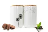 Two white ceramic storage jars with wooden lids, decorated with dandelion motifs, sit on a white surface next to chocolate chip cookies and chocolate pieces with mint leaves. Produktbild-Vorschau