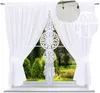 A white voile curtain with a decorative dreamcatcher panel hanging in front of a window with a garden view.