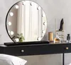 A round vanity mirror with lights sits on a black dresser with some perfume bottles and a decorative shoe. Produktbild-Vorschau