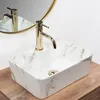 A close-up shot of a rectangular marble-patterned vessel sink with a gold faucet, placed on a wooden surface, with a gold-rimmed mirror reflecting the faucet and sink in the background. Podgląd zdjęcia produktu