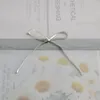 A close-up view of a white ribbon tied in a bow around a light gray band, with a blurred background of white daisies and text. Produktbild-Vorschau