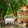 A beige macrame hanging chair with a white cushion is set up on a wooden deck next to a pond, with a rustic wooden house and trees in the background. Produktbild-Vorschau