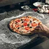 A pizza is being prepared on a floured surface, with ingredients and kitchen equipment in the background. Anteprima dell’immagine prodotto