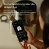 A person is holding a smartphone displaying an app interface, with a kitchen appliance in the background. Produktbild-Vorschau