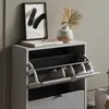 A close-up shot of a gray shoe cabinet with two flip-out drawers, displaying a few pairs of shoes inside. The top surface of the cabinet features a framed abstract artwork, a book, a pair of glasses, and a decorative vase with a dried palm leaf. Podgląd zdjęcia produktu