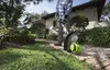 A person is using a Greenworks string trimmer in a garden next to a house. Náhled obrázku produktu