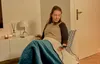 A woman is sitting in a chair wrapped in a blanket, holding a remote control. Produktbild-Vorschau